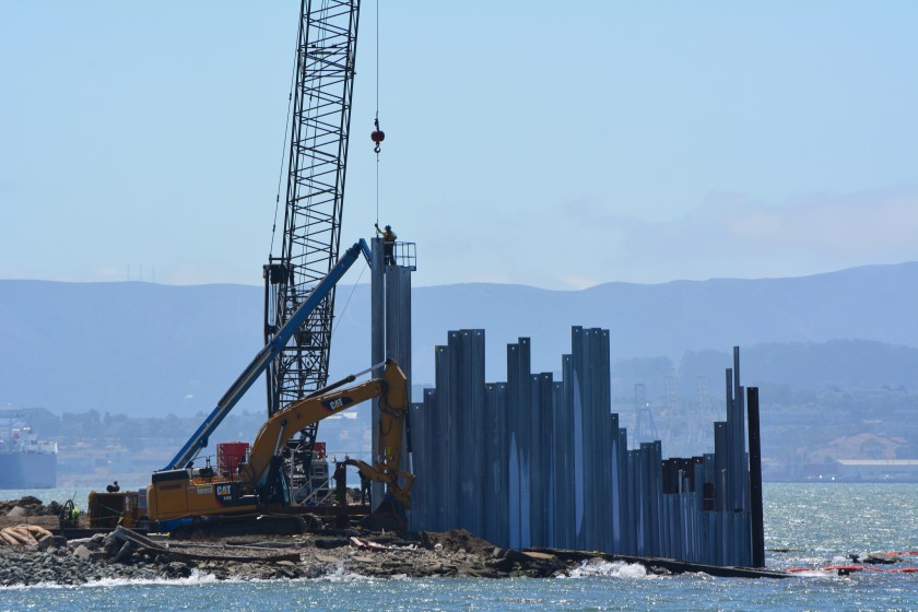 A worker guides one of the galvanized steel pilings into place as crane operator maneuvers.