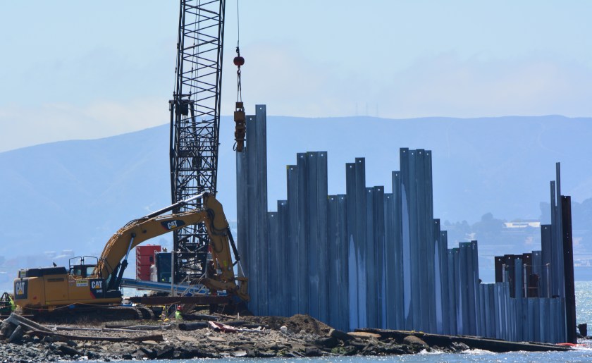 Piling being slowly driven into the ground by a hydraulically operated vibratory hammer.