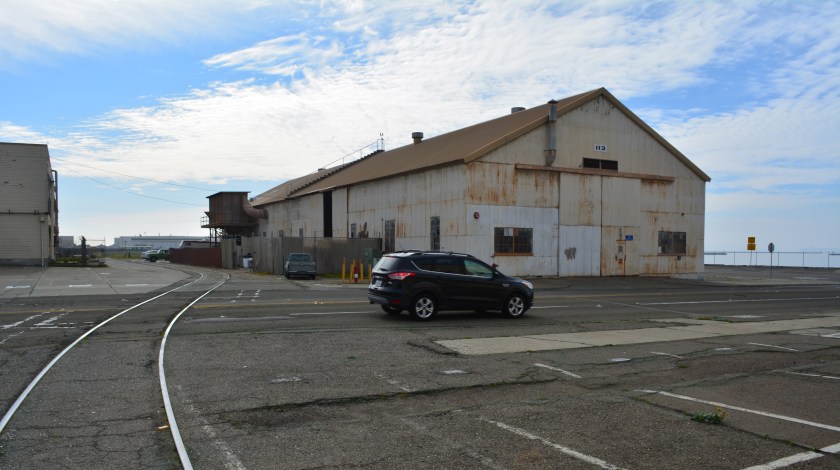 Building 113, which will be reused. Seaplane Lagoon to the right. Ferry Point Road, currently to the right of Building 113, will be realigned to the left of Building 112. W. Atlantic Avenue with car will be moved north to the vantage point of the camera.