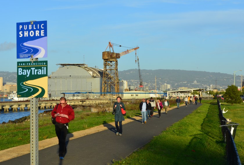 Commuters walking on Bay Trail from ferry terminal to overflow parking lot.  Todd Shipyard Crane and Bay Ship and Yacht floating dry dock in background.  Dog Park on right.