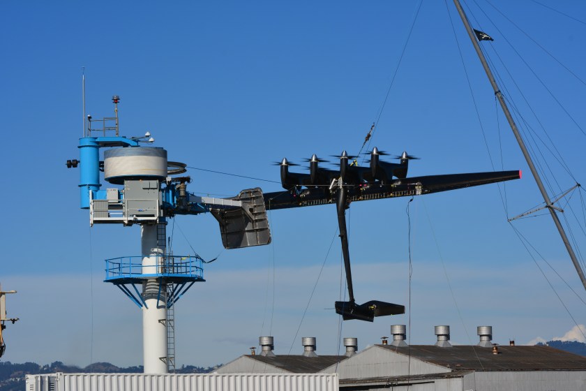 Kite with tether cable extending from tower to belly of kite, with mast suspending the kite during testing. Vertical platform extending from tower is the docking pad for the kite when not flying.