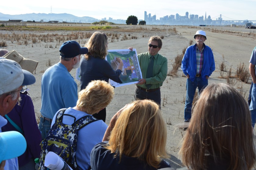 Tour group listening to Jennifer Ott explaining plans of the US Dept. of Veterans Affairs while stopped next to future clinic site. San Francisco skyline in the background.